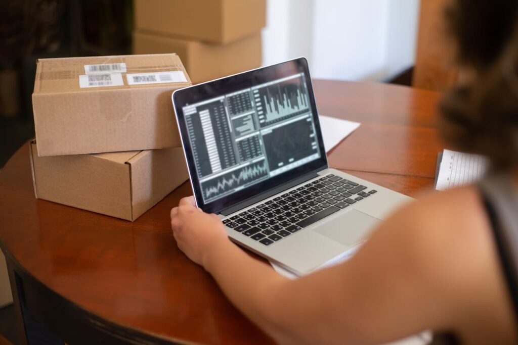 person-looking-at-charts-displayed-on-a-laptop-and-sitting-at-the-table-with-cardboard-boxes-7857526 Person Looking at Charts Displayed on a Laptop and Sitting at the Table with Cardboard Boxes