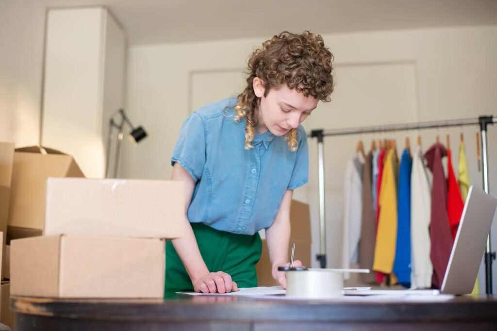 woman-standing-next-to-a-desk-with-a-laptop-and-cardboard-boxes-and-writing-on-paper-7857553 Woman Standing next to a Desk with a Laptop and Cardboard Boxes and Writing on Paper